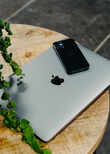 Laptop met een iPhone erbovenop naast een plant liggen op een houten ronde tafel met grijze vloertegels op de achtergrond.