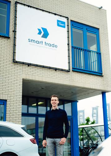 Man stapt uit een witte auto voor het kantoorgebouw van Smart Trade, met het bedrijfslogo zichtbaar op de gevel en een laadpaal op de parkeerplaats.