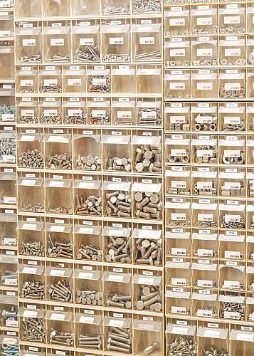 Wand met transparante bakken gevuld met verschillende soorten schroeven, bouten, moeren en ringen, elk voorzien van een label met een artikelnummer.