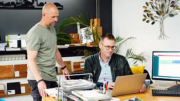 Twee mannen overleggen aan een bureau met Smart Trade software op het scherm, omringd door materialen en groen.