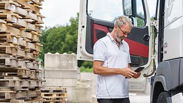 Vrachtwagenchauffeur bekijkt ritplanning op mobiel naast truck, met stapel pallets op de achtergrond.
