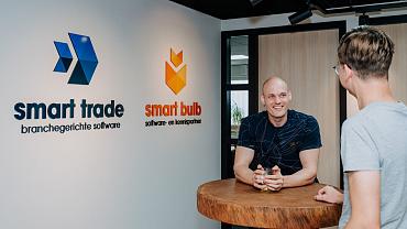 Medewerkers in gesprek aan een hoge tafel op kantoor, met de logo’s van SmartTrade en Smart Bulb zichtbaar op de muur op de achtergrond.