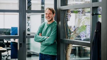 Medewerker van Smart Trade staat glimlachend met gekruiste armen in een moderne kantoorruimte met glazen wanden en een plant met paarse bloemen op de achtergrond.