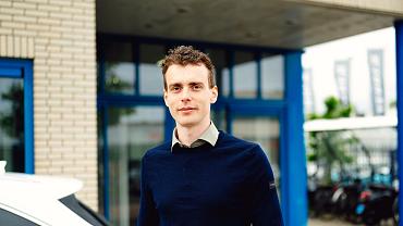 Man in donkerblauwe trui staat buiten bij bedrijfsgebouw met blauwe kozijnen, naast geparkeerde auto.