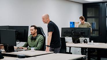 Drie collega’s werken samen op kantoor aan meerdere beeldschermen, terwijl één medewerker meekijkt en ondersteuning geeft bij een taak op de computer.