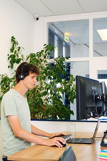 Man staat aan bureau met koptelefoon op te werken op zijn computer. Op de achtergrond staat een grote plant en hangt een scherm met data.